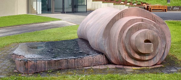 Skulptur aus Stein vor einem modernen Gebäude mit Glas und Grünfläche